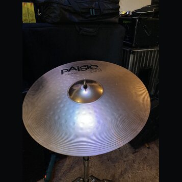 Stříbrný cymbál s koncentrickým žebrováním, umístěný na kovovém stojanu. Na povrchu je černý nápis PAiste 2008 bronze a pod ním text 18" CRASH RIDE.…
