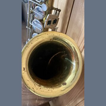 Detailní pohled na čňel mosazného saxofonu. Povrch uvnitř i vně čněle vykazuje výrazné známky opotřebení, oxidaci a tmavé skvrny. V horní části je vi…