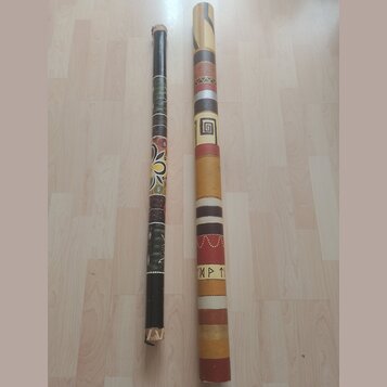 Dvě didgeridou z dřeva s různým povrchovým zdobením. Levý nástroj je tmavě hnědý s barevnými ornamenty a vzory. Pravý nástroj je světlejší, zdobený h…