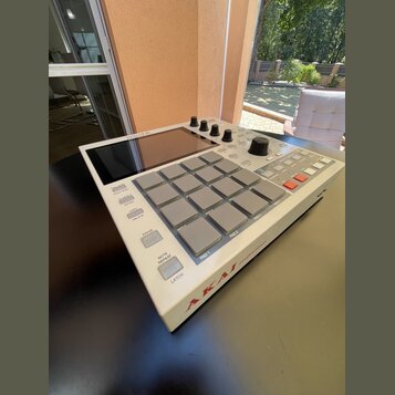 Bíle provedený sampler s mřížkou 16 šedých padů a displejem v horní části. Na spodní hraně je červený nápis AKAI a vedle něj nečitelný text. Zařízení…
