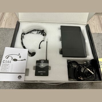 V bílé krabici s polystyrenovými vložkami je černý bezdrátový mikrofonní set. Sada obsahuje náhlavní mikrofon, vysílač s nápisy AKG, PERCEPTION wirel…