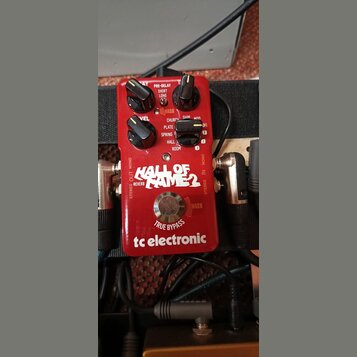 Červený efekt pedál s nápisy HALL OF FAME 2, REVERB a tc electronic. Na horní části jsou čtyři černé ovládací knoflíky s popisky PRE-DELAY, LEVEL a d…