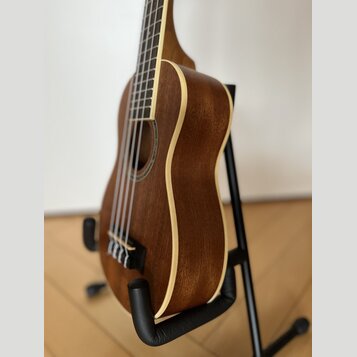 Hnědý akustický nástroj typu ukulele s dřevěným povrchem a světlým lemováním. Nástroj je umístěn v černém stojanu. Na viditelné části nejsou žádné zn…