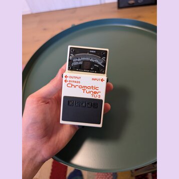 Bílý efekt pedál s černým spínacím pedálem a analogovým ukazatelem v horní části. Na zařízení jsou nápisy BOSS, Chromatic Tuner TU-3 a označení vstup…