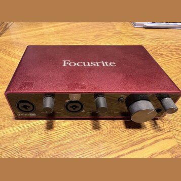 Červená audio rozhraní s nápisem Focusrite na horní straně. Na předním panelu jsou dva kombinované vstupy, dva ovládací knofíky zyska a velký knofík…