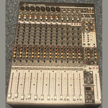 Šedý mixovací pult s mnoha ovládacími prvky, posuvníky a konektory. V horní části je nápis Behringer XENYX UFX1604. Povrch přístroje vykazuje mírné z…