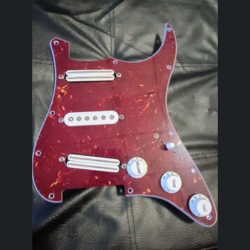 Vinylový pickguard v tmavě červené barvě s texturou želvy, osazený třemi bílými snímači. Na ovládacích knoflících jsou nápisy VOLUME, TOINE a ONE. So…