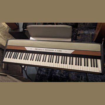 Digitální piano s klávesnicí, kombinací dřevěného a šedého povrchu. Na ovládacím panelu je vlevo nápis KORG a vpravo označení modelu SP-250. Nad kláv…