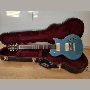 Modře lakovaná elektrická kytara typu Les Paul v černém tvrdém kufru s červeným polstrováním. Na hlavě hmatníku je bílý nápis Summit a text MADE IN K…