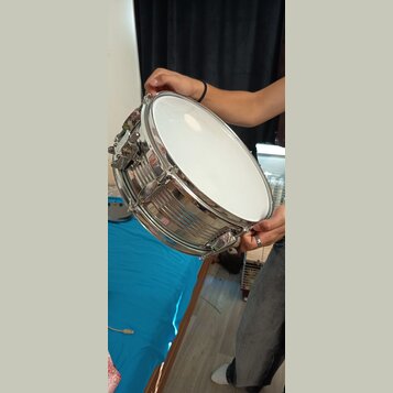 Fotografie zachycuje snare drum s chromovaným kovovým korpusem a bílým membránou. Nástroj drží osoba v rukou. Na viditelných částech nástroje nejsou…