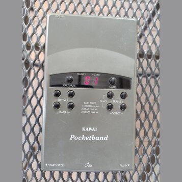 Šedý plastový přístroj s nápisy KAWAI a Pocketband. Na přední straně je digitální displej s červenými číslicemi a několik ovládacích tlačítek. Jsou z…