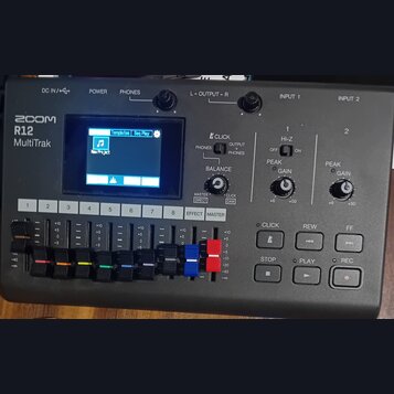 Černý digitální záznamový zařízení s nápisem ZOOM R12 MultiTrak. Přístroj má v horní části displej, dva regulační knofíky pro výstup a dvě sekce vstu…