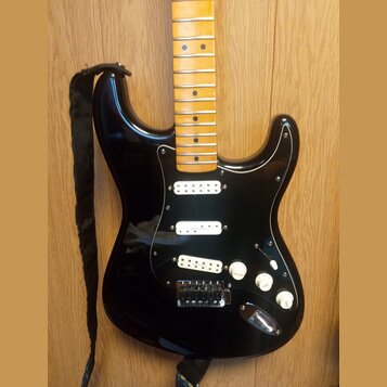 Černá elektrická kytara s tvarem typu Stratocaster a černým pickguardem. Má světle hnědý javorový hmatník a tři bílé snímače. Na pickguardu je vidite…