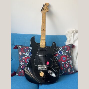 Černá elektrická kytara s mírně prohnutým tělem a světle hnědým hnedem hmatníkem. Na hlavě je nápis Squier Stratocaster. Tělo má černý pickguard, tři…