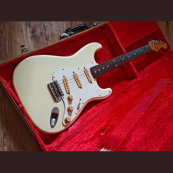 Bílá elektrická kytara typu Stratocaster s bílým pickguardem a hnedým hmatníkem, umístěná v otevřeném kufru s červeným polstrováním. Na hlavě hmatník…