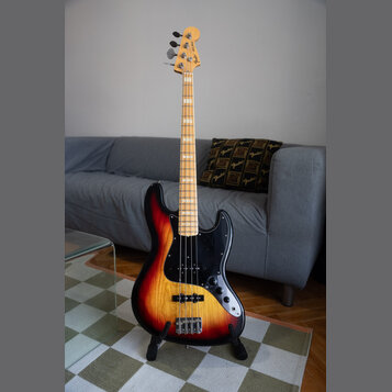 Elektrická basová kytara s trupem v barevném přechodu od žluté přes oranžovou k červené a černé. Nástroj má černý pickguard, javorový hmatník a světl…