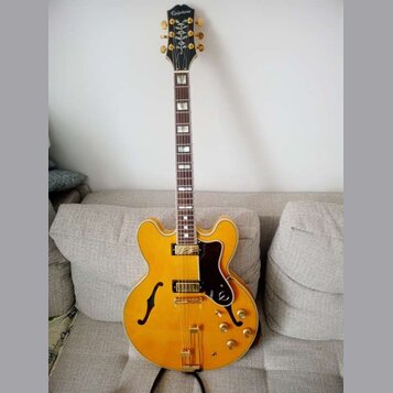 Žlutá polodupatá elektrická kytara s hnědým hmatníkem a černým pickguardem. Na hlavě je bílý nápis Epiphone a dekorativní ornament. Nástroj má zlaté…