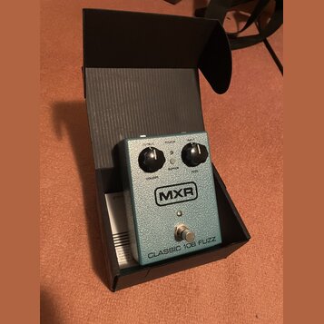 Krytá krabička s efekt pedálem v modro-zelené barvě s texturovaným povrchem. Na pedálu je logo MXR a nápis CLASSIC 108 FUZZ. Jsou zde dva černé ovlád…
