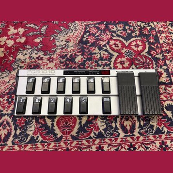 Stříbrný MIDI pedál s deseti černými spínači a dvěma expresními pedály v dolní části. Na pravém boku je svisle napsáno FCB1010 MIDI FOOT CONTROLLER.…