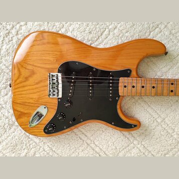 Elektrická kytara s přírodním dřevěným povrchem a černým pickguardem. Nástroj má tři snímače typu single-coil, jeden mostový snímač typu humbucker a…