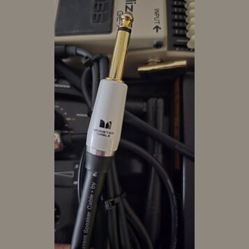 Fotografie zachycuje konektor typu jack v zlatém provedení s bílým plastovým krytem. Na krytu je černé logo ve tvaru písmene M a nápis MONSTER CABLE.…