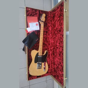 Elektrická kytara typu Telecaster v přírodní dřevěné barvě s černým pickguardem, umístěná v otevřeném kufru s červeným plyšovým vnitřkem. Na hlavě hm…