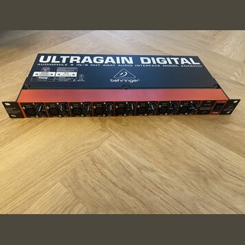 Černý rackový audio interface s červeným horním panelem. Na spodní straně je nápis ULTRAGAIN DIGITAL AUDIOPHILE 8 IN/8 OUT ADAT AUDIO INTERFACE MODEL…