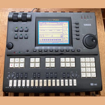Šedý music sequencer značky YAMAHA, model QY700. Zařízení má v horní části displej s modrým podsvícením a pod ním řadu ovládacích tlačítek. V pravé č…