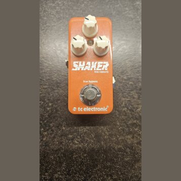 Oranžový efekt pedál s třemi bílými oválnými knoflíky. Na přední straně je bílý nápis SHAKER a pod ním menší text MINI VIBRATO. Pod ním je uveden náp…