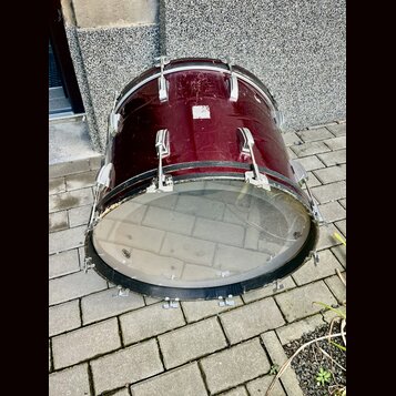 Basový buben v tmavě červené barvě s kovovým kováním. Povrch nástroje vykazuje viditelné opotřebení, škrábance a několik světlých skvrn. Na přední st…