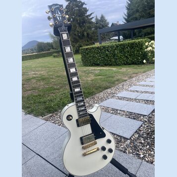 Bílá elektrická kytara s černým hmatníkem a černým pickguardem, umístěná na stojanu. Na hlavě hmatníku je zlaté logo Gibson. Nástroj má dva humbucker…