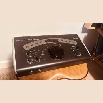 Černý monitorovací kontrolér s bílým rámem a nápisem BEHRINGER v pravém horním rohu. Na předním panelu je uveden model XENYX CONTROL 2 USB a jsou zde…