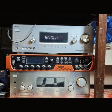 Tři stohované elektronické komponenty v regálu. Horní je stříbrný receiver s nápisy SONY, Digital Audio/Video Control Center a HDMI. Prostřední je or…