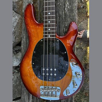 Elektrická basová kytara s hnědým slunečním odstínem (sunburst) a černým kruhovým pickguardem. Na horní části těla je vidět nápis Rickenbacker a chro…