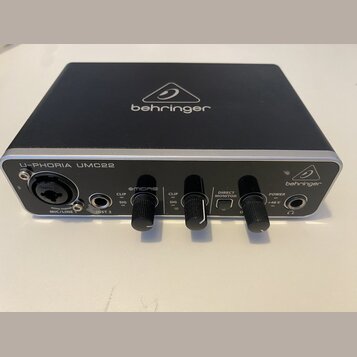 Černý audio rozhraní s hliníkovým stříbrným horním panelem. Na přední straně je nápis U-PHORIA UMC22 a logo behringer, stejně jako na horní části zař…
