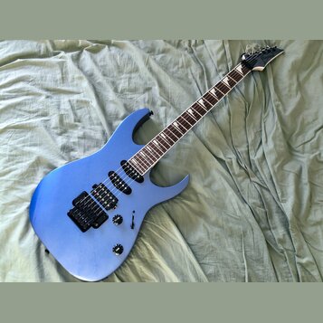 Modrá elektrická kytara s tmavým hmatníkem a černými komponenty. Na hlavě krku je bílý nápis Ibanez. Nástroj má dva humbuckery, jeden ovládací knoflí…