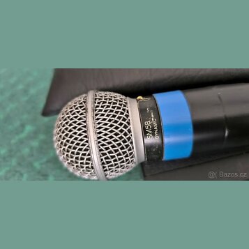 Detailní pohled na dynamický mikrofon s černým tělem a modrým kroužkem. Na stříbrném prstenci pod mřížkou je černý nápis SM58 DYNAMIC. Kovová mřížka…