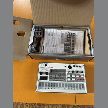 Na fotografii je bílý sampler KORG volca sample v originální krabici. Za přístrojem je otevřený kartonový obal, ve kterém se nachází uživatelský manu…