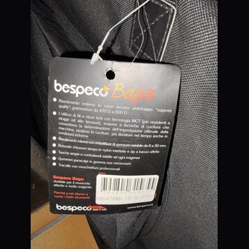 Fotografie detailu černé textilní tašky s připevněným černým papírovým štítkem. Na štítku je nápis bespeco Bags a text v italštině popisující vlastno…