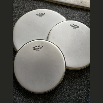 Tři bílé kruhové bubnové blány s kovovými obručemi. Každá blána má v horní části černé logo REMO a pod ním nápis EMPEROR® WHITE COATED MADE IN U.S.A.…