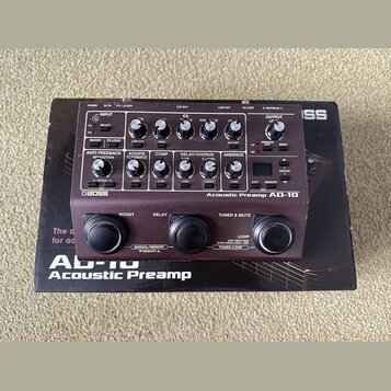 Černý kytarový pedál s nápisy BOSS a Acoustic Preamp AD-10. Zařízení má na horním panelu řadu ovládacích knoflíků, přepínačů a malý digitální displej…