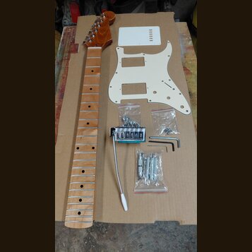 Sada komponentů pro elektrickou kytaru položená na hnědé kartonové ploše. Obsahuje dřevěný krk s hnědým lakem, bílý plastový pickguard a bílou krytku…