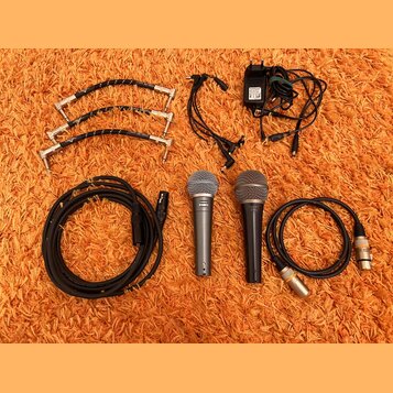 Sada vybavení složená ze dvou dynamických mikrofonů, kabelů a napájecího adaptéru. Na mikrofonech jsou viditelné nápisy "Sennheiser" a "Cobalt 810".…