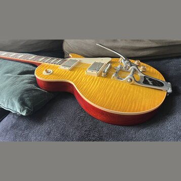 Elektrická kytara s žlutým plamenovitým topem a červenými boky. Na kytaru je instalován chromovaný Bigsby vibrační systém s nápisem „Bigsby“. Nástroj…