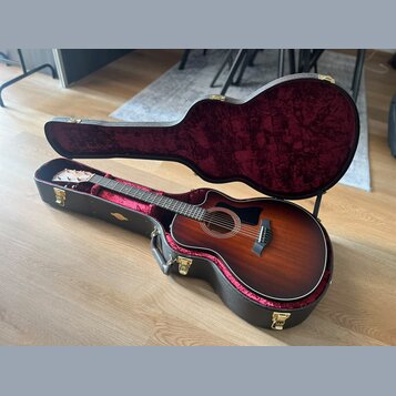Akustická kytara s cutawayem v tmavě hnědém sunburst provedení, umístěná v otevřeném černém tvrdém pouzdře s červeným plyšovým vnitřkem. Na hlavě hma…
