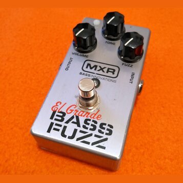 Stříbrný efekt pedál s třemi černými ovládacími knoflíky označenými jako VOLUME, TONE a FUZZ. Na horní části je logo MXR a nápis BASS INNOVATIONS, ve…