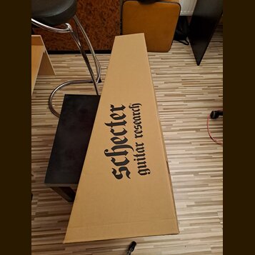 Kartonový obal v hnědé barvě pro hudební nástroj. Na povrchu je černý nápis Schecter guitar research. Obal vykazuje mírné pomačkání v dolní části.