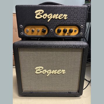 Kytarový zesilovač a reproduktorové boxy značky Bogner. Horní jednotka má zlatý ovládací panel s nápisy BARCELONA, volume, gain, bass a treble. Předn…