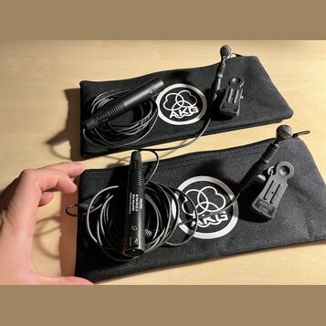 Dvě černé zipové pouzdra s bílým logem AKG. V každém pouzdře je černý mikrofon s připojeným kabelem a držákem. Na spodním mikrofonu je nápis PHANTOM…