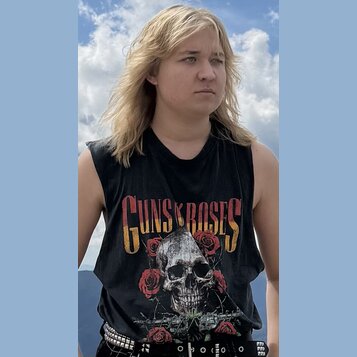 Fotografie zobrazuje osobu v černém tričku bez rukávů. Na přední straně trička je žlutý nápis GUNS N ROSES a grafika lebky s červenými růžemi. Osoba…